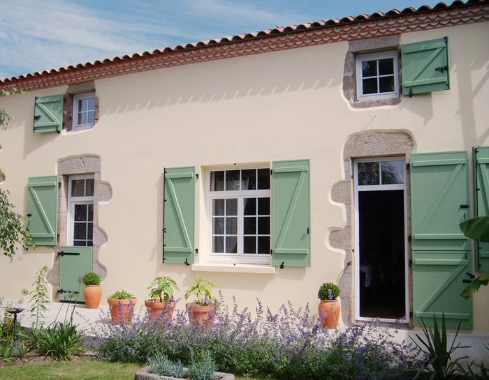 Façade beige, fenêtres blanches encadrées pierre, volets bois vert sauge, pots terre cuite