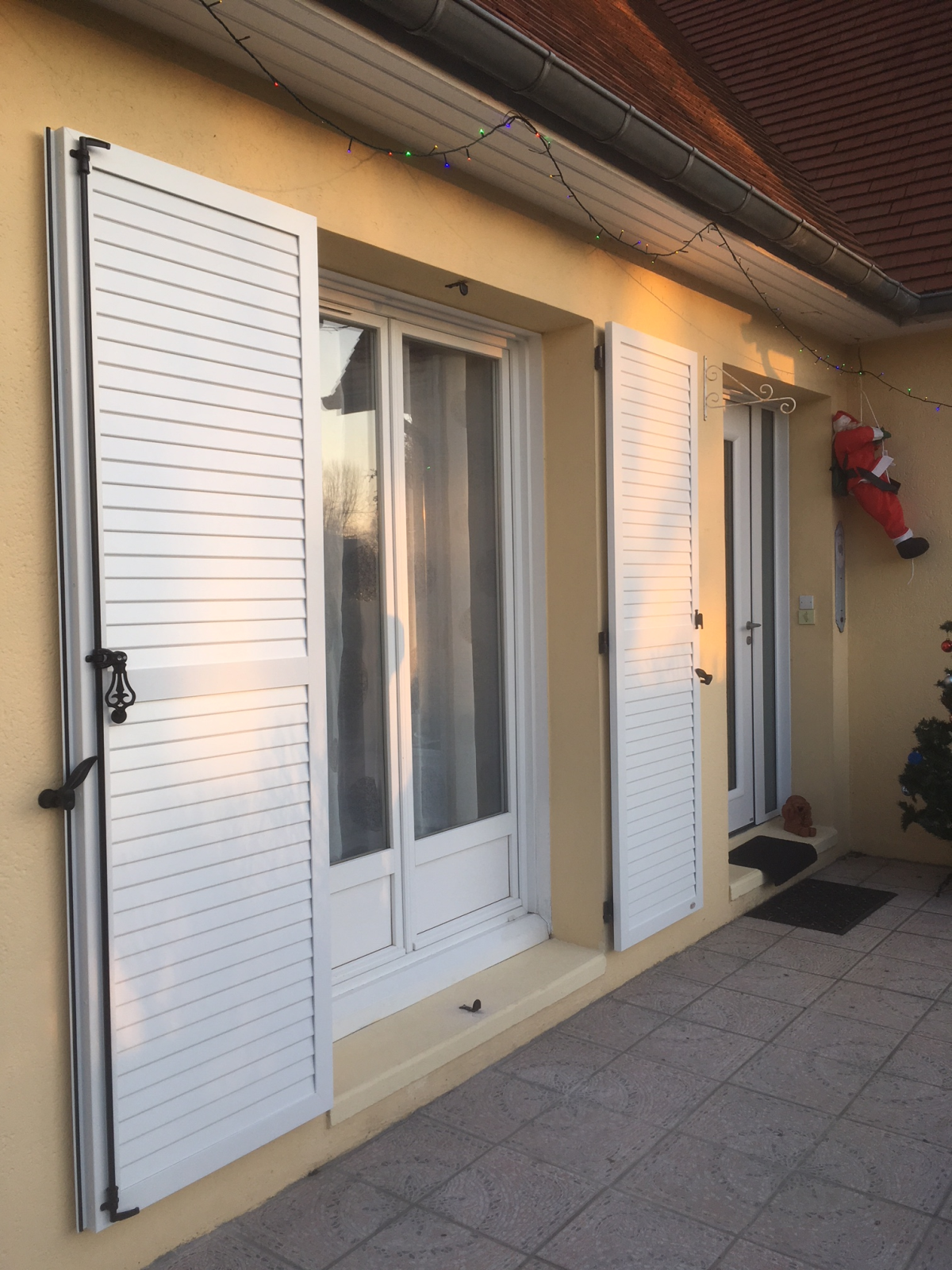 Volets battants blancs à persiennes et ferrures noires autour d’une porte-fenêtre, façade jaune