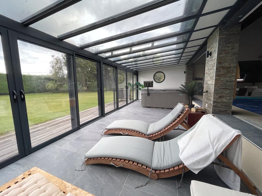 Véranda vitrée toiture inclinée gris anthracite, spa gris et fauteuils bois sur carrelage
