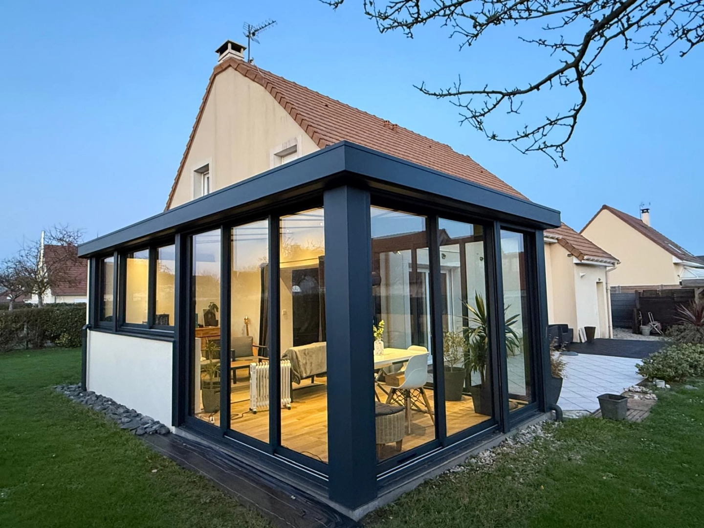 Véranda gris anthracite vitrée sur maison claire, intérieur éclairé au crépuscule
