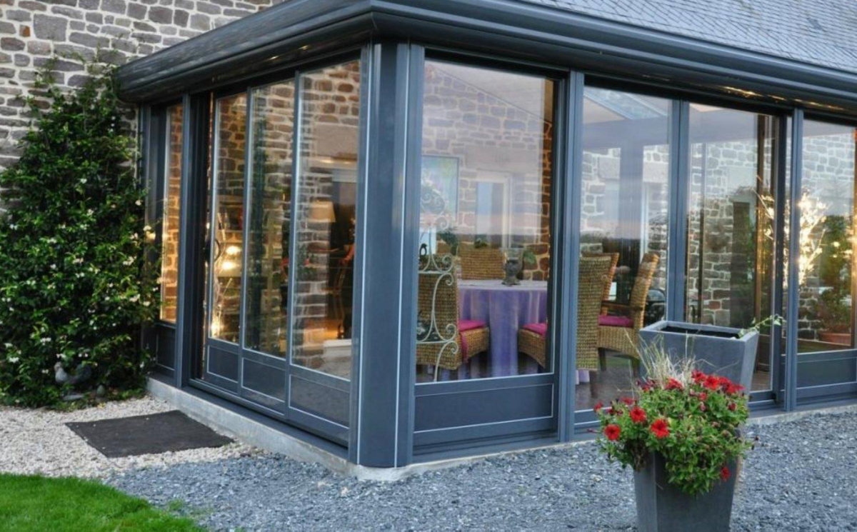 Véranda gris anthracite vitrée sur maison pierre, table violette et chaises osier éclairées