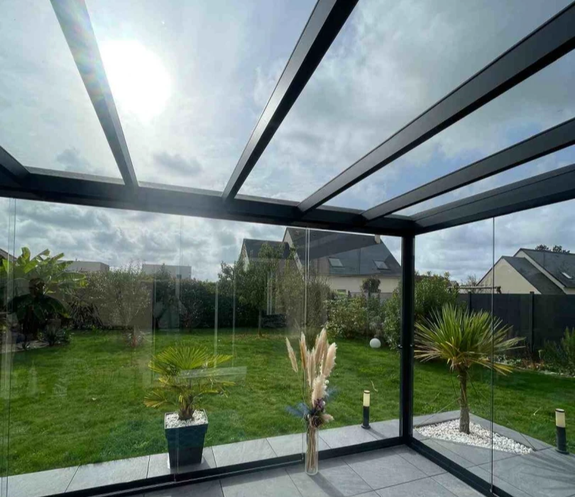 Véranda vitrée cadre noir, carrelage gris, fauteuils courbes et jardin vu par les vitres