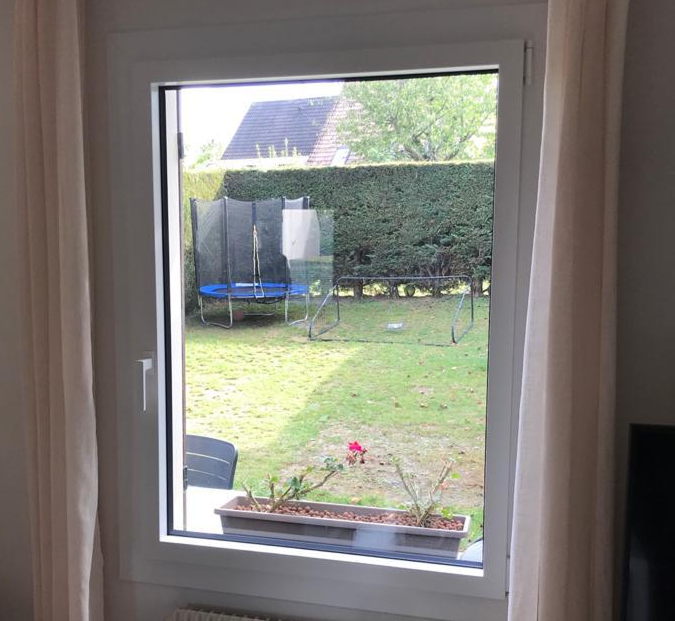 Fenêtre PVC blanche intérieure, rideaux crème, radiateur et vue jardin trampoline
