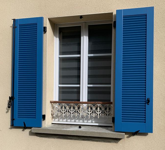 Volets battants à lames orientables bleus sur une façade de maison beige crème