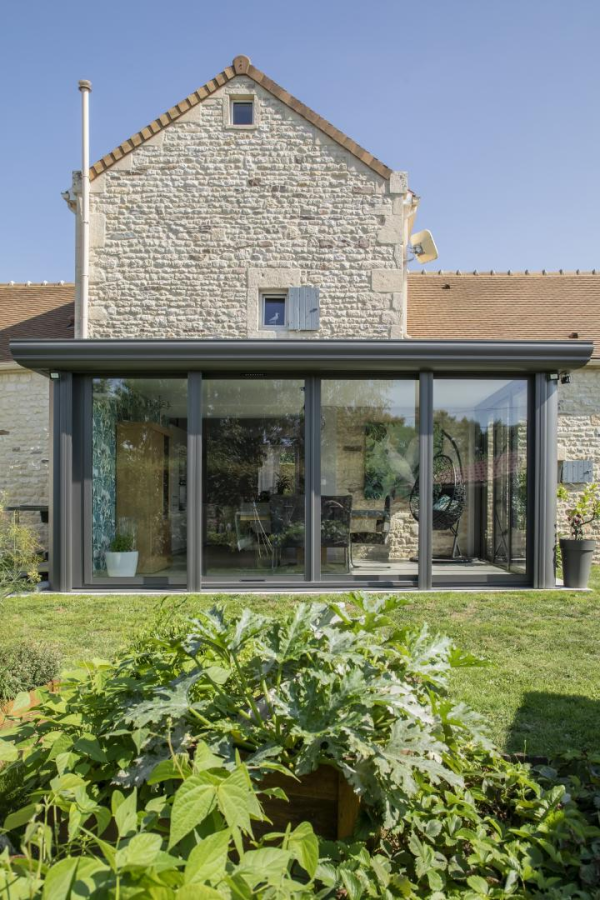 Extension vitrée gris anthracite sur maison pierre, fauteuil œuf noir et potager devant