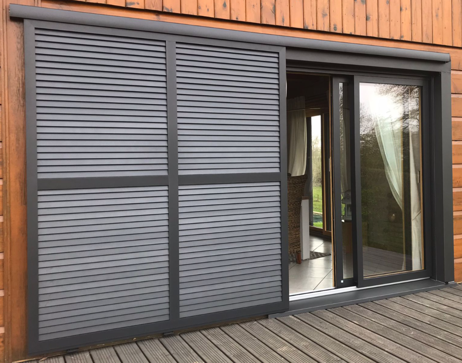 Volet coulissant anthracite à lames sur baie d’un chalet bois, terrasse et campagne verdoyante