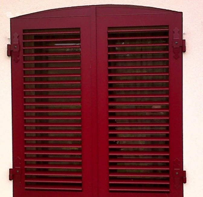 Volets battants persiennes rouge bordeaux cintrés, pentures noires, mur crépi beige