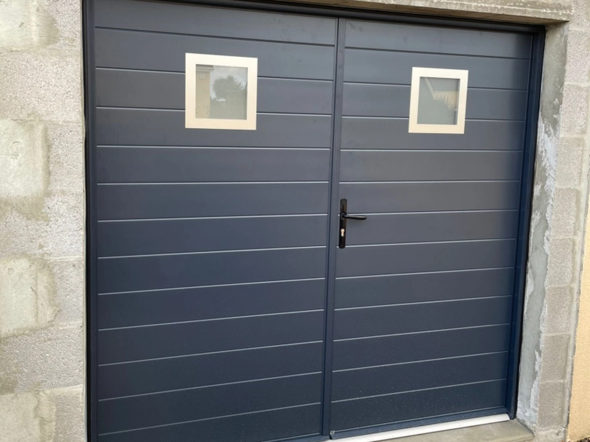 Porte de garage battante anthracite à rainures et hublots carrés dans mur parpaings gris