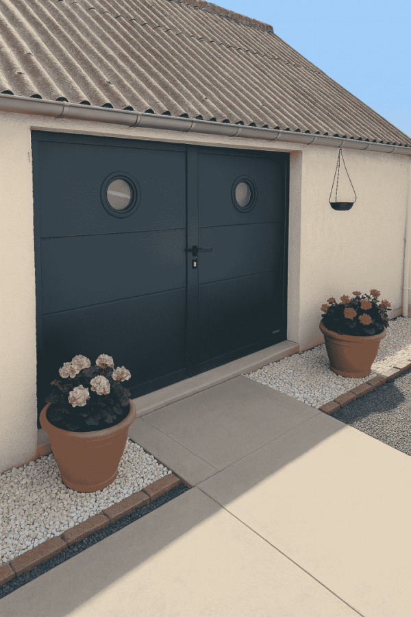 Porte garage battante anthracite deux vantaux, hublots ronds, terrasse béton et graviers