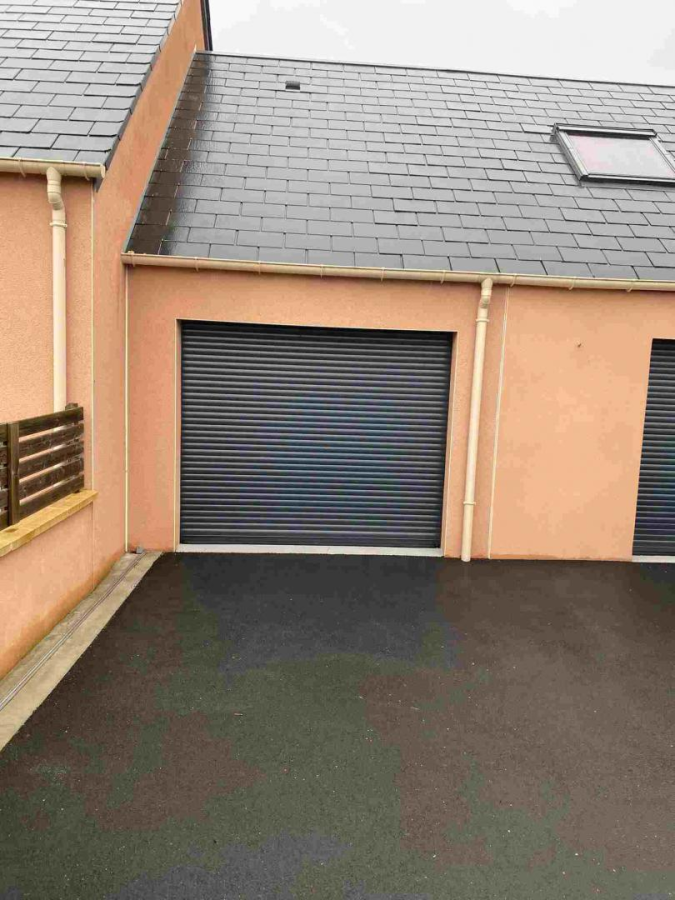 Porte garage enroulable anthracite, façade saumon, toit ardoise, allée bitume mouillée