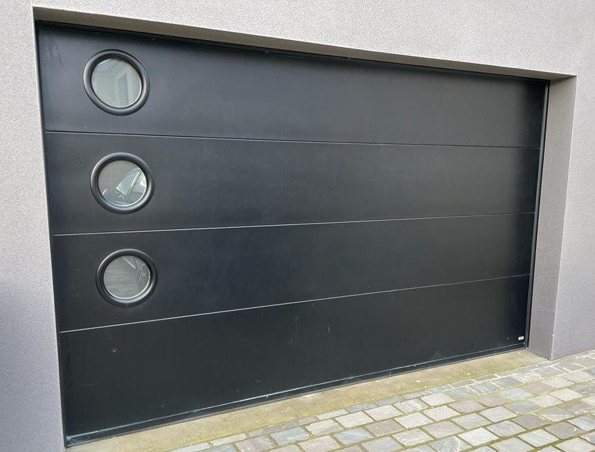 Porte garage sectionnelle noire, trois hublots ronds à gauche, mur crépi gris, pavés