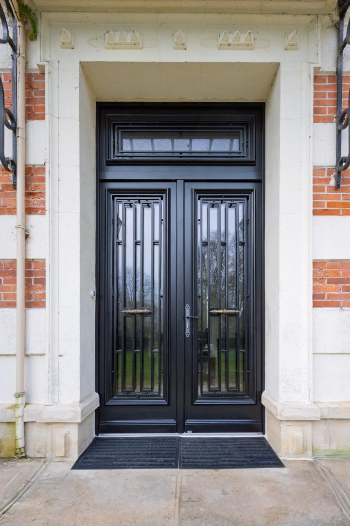 Double porte noire vitrée, grilles art déco, imposte, encadrement pierre et bandeaux briques blanches
