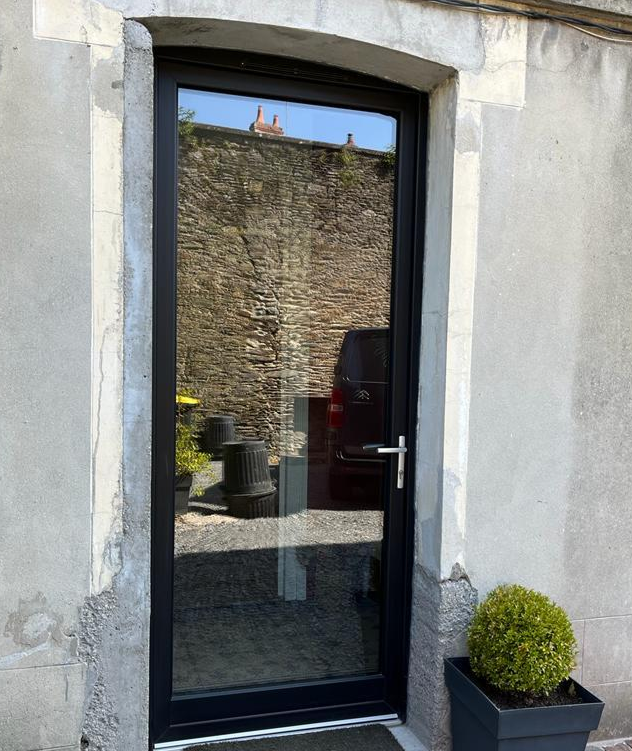 Porte vitree cadre noir dans mur en pierre, paillasson gris et arbuste en pot