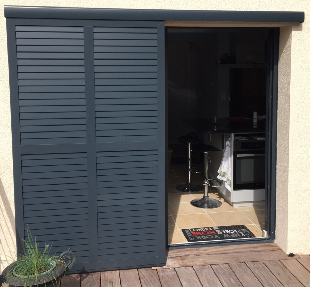 Volet coulissant anthracite à lames persiennes sur terrasse bois, ouverture vers cuisine moderne