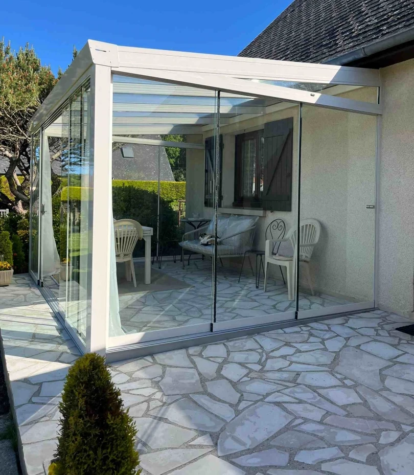 Veranda blanche a parois vitrees coulissantes sur terrasse pierre et volets marron