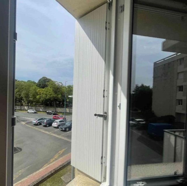 Fenêtre PVC blanche ouverte, volet battant à lames verticales plié, vue parking arboré
