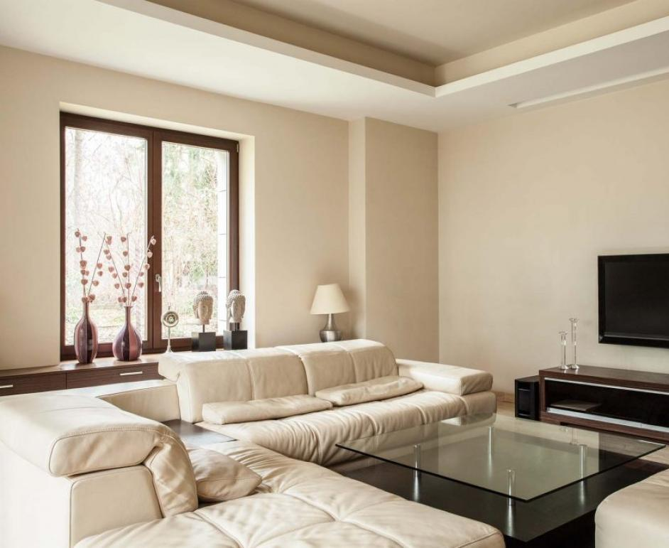 Grand salon avec fenêtre cadre brun, canapé blanc en L et table basse vitrée, vue sur arbres