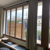 Grande fenêtre bois clair multi-vitrages et rideaux beiges, vue intérieure sur jardin clôturé