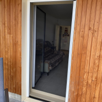 Moustiquaire sur ouverture cadre beige, bardage bois vertical clair, intérieur carrelé visible