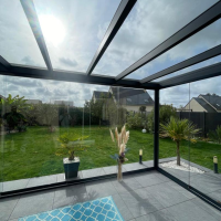 Véranda alu noir vitrée, porte coulissante ouverte sur jardin avec pelouse et palmiers