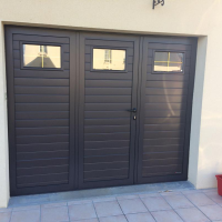 Porte garage gris anthracite trois vantaux, lames horizontales et hublots rectangulaires sur façade crème