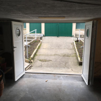 Porte battante blanche double ouverte, hublots ronds, vue intérieur vers rampe béton et portail vert