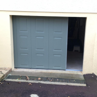Porte garage coulissante gris bleu entrouverte, panneaux à moulures sur mur beige et seuil béton