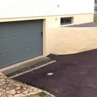 Porte garage latérale grise à embossages rectangulaires, façade beige et fenêtre sous-sol à barreaux