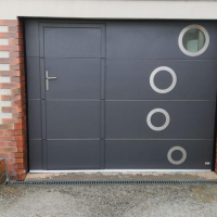 Porte sectionnelle anthracite avec portillon, hublot rond et anneaux décoratifs argent, mur brique