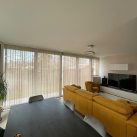 Stores verticaux beige clair sur grandes baies dans salon moderne, canapé moutarde et sol clair