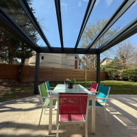 Pergola anthracite toiture vitrée, table blanche et chaises colorées sur terrasse claire