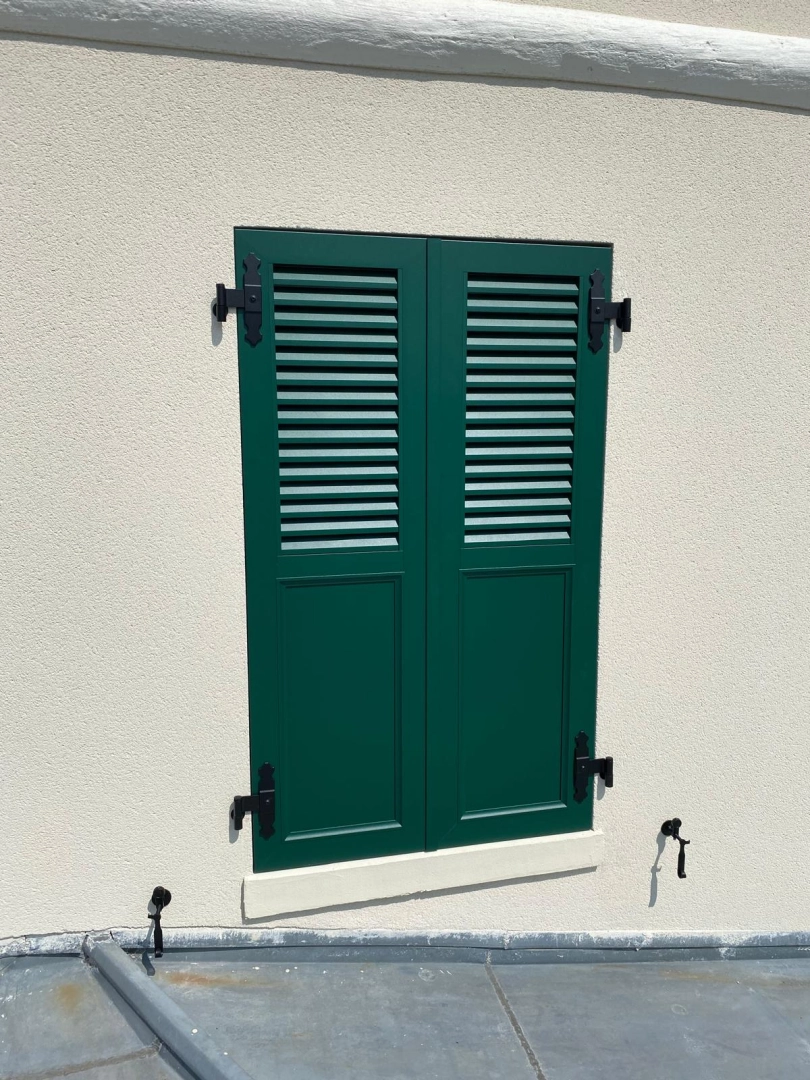 Volets battants vert sapin, persiennes en partie haute et panneaux pleins en bas sur façade beige