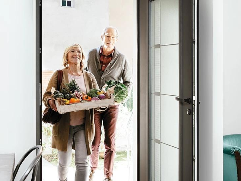 Porte d'entrée aluminium anthracite ouverte, couple portant une caisse de légumes