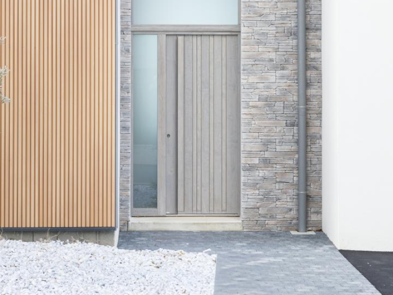 Porte d'entrée bois gris clair avec vitrage latéral et imposte, mur pierre