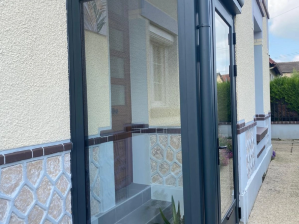Sas d'entrée alu anthracite vitré devant porte bois, façade crépi beige et pierre bleutée
