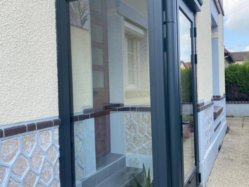 Sas d'entrée alu anthracite vitré devant porte bois, façade crépi beige et pierre bleutée
