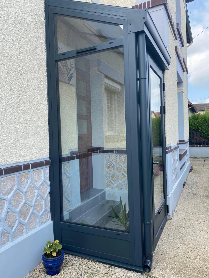Sas d'entrée alu anthracite vitré devant porte bois, façade crépi beige et pierre bleutée