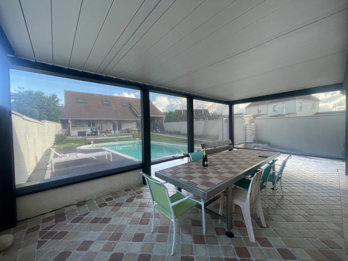 Pergola bioclimatique fermée par parois vitrées, table en pierre, piscine visible au fond