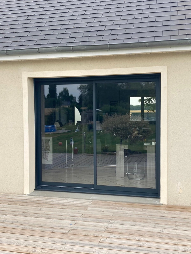 Baie coulissante alu anthracite sur façade enduit beige, toiture ardoises grises