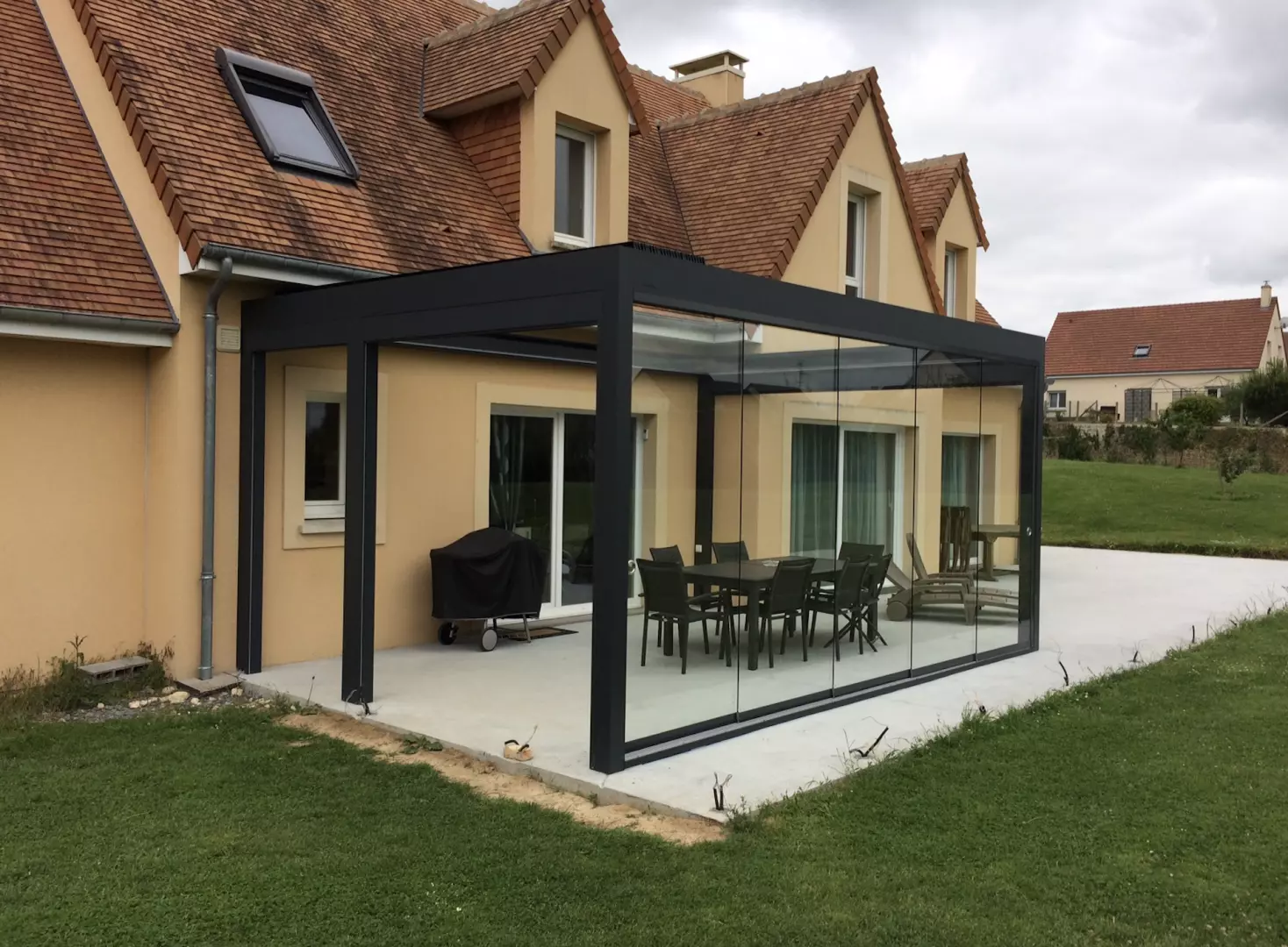 Véranda alu noir toit plat, parois vitrées coulissantes, dalle béton, salon et barbecue