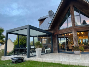 Pergola Brustor au crepuscule, lames blanches, verre et maison bardage bois