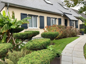 Façade beige, trois fenêtres stores zébrés et volets anthracite, jardin taillé