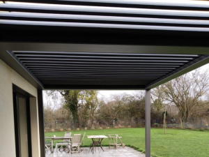Sous pergola grise, dalles claires, table bois et pelouse avec arbres dénudés