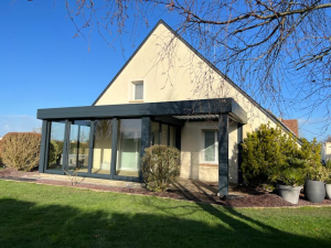 Extension anthracite vitrée et pergola sur pavillon beige, pelouse et arbustes