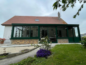 Veranda vitree vert fonce et briques claires le long d'une maison crepi sous ciel gris