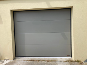 Face porte sectionnelle grise mate, seuil aluminium et herbes au pied du mur beige