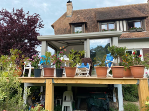 Extension vitree grise sur terrasse bois sur pilotis, pots fleuris devant maison tuiles