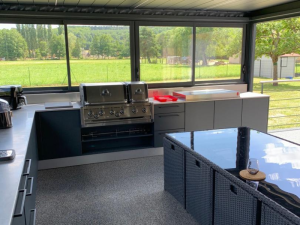 Cuisine extérieure inox sous pergola, grill encastré et baie vitrée sur prairie