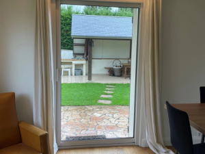 Porte-fenetre blanche, rideaux creme et vue terrasse pierres et pelouse verte