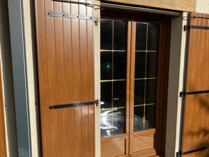 Volets effet bois ouverts, porte vitrée croisillons dorés sur crépi beige